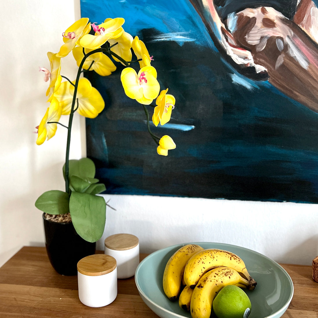 A realistic artificial yellow orchid with a pink center and green leaves in a ceramic vase, displayed on a table with bananas and a bowl in the foreground.